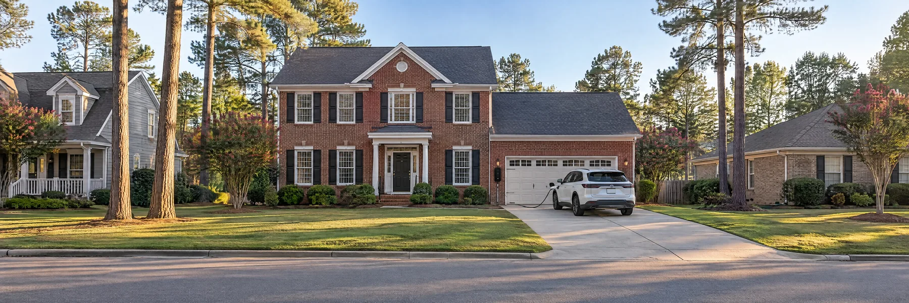 Red brick two-story house with a white SUV in the driveway, plugged into a charger beside the garage door, under tall pines
